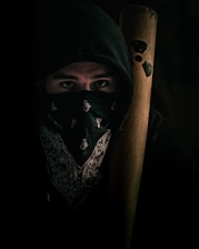 A person wearing a dark hoodie and a patterned bandana over the lower part of their face. They hold a wooden bat adorned with a radiation symbol. The lighting is low, creating a mysterious and tense atmosphere.