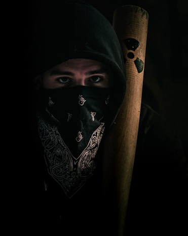 A person wearing a dark hoodie and a patterned bandana over the lower part of their face. They hold a wooden bat adorned with a radiation symbol. The lighting is low, creating a mysterious and tense atmosphere.