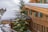 A wooden chalet with green shutters on the windows is covered by a thick layer of snow on the roof. Tall evergreen trees are partially covered in snow, surrounding the chalet, with a mountainous backdrop partially visible. Another similar chalet is seen in the distance.