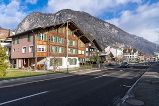 A picturesque street scene in a mountainous region featuring a traditional wooden chalet-style building with green shutters. The building is surrounded by a small garden and sits next to a road with a few cars. In the background, there are snow-capped mountains under a partly cloudy sky.