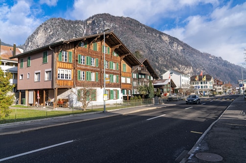 A picturesque street scene in a mountainous region featuring a traditional wooden chalet-style building with green shutters. The building is surrounded by a small garden and sits next to a road with a few cars. In the background, there are snow-capped mountains under a partly cloudy sky.