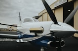 A small, sleek propeller airplane is positioned outside a hangar. The aircraft's polished body reflects light, and its propeller is prominently displayed in the foreground. The hangar behind features a sign with partial text visible. The scene conveys a sense of readiness and adventure.