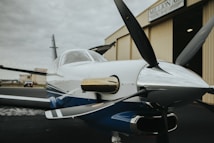 A small, sleek propeller airplane is positioned outside a hangar. The aircraft's polished body reflects light, and its propeller is prominently displayed in the foreground. The hangar behind features a sign with partial text visible. The scene conveys a sense of readiness and adventure.
