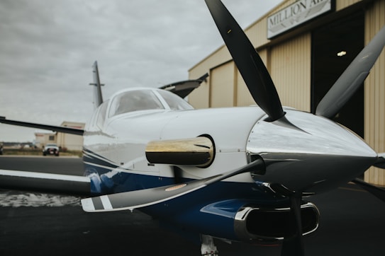 A small, sleek propeller airplane is positioned outside a hangar. The aircraft's polished body reflects light, and its propeller is prominently displayed in the foreground. The hangar behind features a sign with partial text visible. The scene conveys a sense of readiness and adventure.