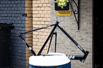 A black bicycle frame is displayed on a barrel covered in snow. It is positioned against a backdrop of brick walls painted in different shades, one in gray and another in a tan color. A sign with a speech bubble design hangs on the wall nearby, indicating business hours and a phone number. Snowflakes are visibly falling in the scene.