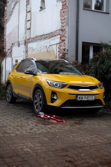 A bright yellow car is parked on a cobblestone surface near a partially renovated building. The car is a modern SUV with a visible European license plate. In front of the car, two folded red and white warning signs are placed on the ground. The surrounding area includes exposed brickwork and unfinished construction, with climbing ivy on the side.