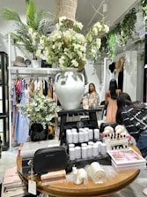 A boutique store interior with a prominent display table featuring a large white vase filled with lush white flowers. The table also showcases candles, books, and small decorative items. Surrounding the table are racks with diverse clothing, a woman browsing, and lush greenery providing a fresh ambiance.
