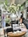 A boutique store interior with a prominent display table featuring a large white vase filled with lush white flowers. The table also showcases candles, books, and small decorative items. Surrounding the table are racks with diverse clothing, a woman browsing, and lush greenery providing a fresh ambiance.