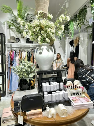 A boutique store interior with a prominent display table featuring a large white vase filled with lush white flowers. The table also showcases candles, books, and small decorative items. Surrounding the table are racks with diverse clothing, a woman browsing, and lush greenery providing a fresh ambiance.
