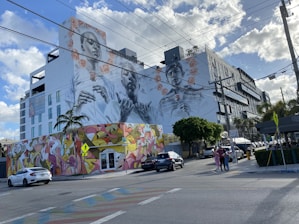 A large mural on the side of a building features intricately painted portraits of two people. The mural is adorned with floral patterns, and below it, the building's lower section is covered with colorful abstract graffiti. Cars are parked and driving on the street, with scattered pedestrians walking nearby. Overhead, power lines stretch across the scene against a partly cloudy sky.