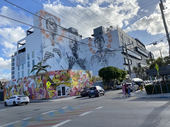 A large mural on the side of a building features intricately painted portraits of two people. The mural is adorned with floral patterns, and below it, the building's lower section is covered with colorful abstract graffiti. Cars are parked and driving on the street, with scattered pedestrians walking nearby. Overhead, power lines stretch across the scene against a partly cloudy sky.