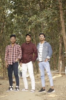 Three young men stand on a forest path surrounded by tall trees. Each displays a different style: one wears a plaid shirt and jeans with sneakers, another sports a maroon jacket and white pants with sandals, and the third has on a denim jacket and striped shirt with distressed jeans and sneakers. They are posing casually, their expressions relaxed.