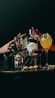 Several bottles and glasses of alcoholic beverages are arranged on a dimly lit table. Hands with painted nails are reaching for the bottles, and the drinks are garnished with fruits and leaves. The scene is set against a dark background, suggesting a bar or party atmosphere.