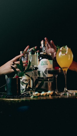 Several bottles and glasses of alcoholic beverages are arranged on a dimly lit table. Hands with painted nails are reaching for the bottles, and the drinks are garnished with fruits and leaves. The scene is set against a dark background, suggesting a bar or party atmosphere.