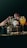 Several bottles and glasses of alcoholic beverages are arranged on a dimly lit table. Hands with painted nails are reaching for the bottles, and the drinks are garnished with fruits and leaves. The scene is set against a dark background, suggesting a bar or party atmosphere.