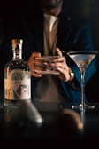 A person is shuffling a deck of playing cards next to a bottle of gin and a martini glass with an olive. The lighting is dim, creating a mysterious ambiance.