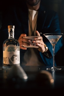 A person is shuffling a deck of playing cards next to a bottle of gin and a martini glass with an olive. The lighting is dim, creating a mysterious ambiance.