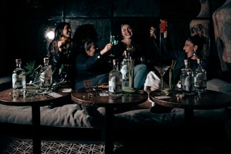 A group of people are sitting together on a sofa, raising glasses in a celebratory manner. The setting appears to be a dimly lit, cozy room with a relaxed atmosphere. Three tables in the foreground display bottles of liquor, playing cards, and some green plants.