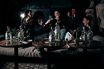 A group of people are sitting together on a sofa, raising glasses in a celebratory manner. The setting appears to be a dimly lit, cozy room with a relaxed atmosphere. Three tables in the foreground display bottles of liquor, playing cards, and some green plants.
