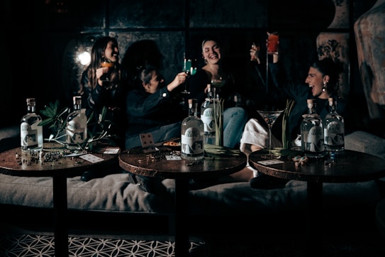 A group of people are sitting together on a sofa, raising glasses in a celebratory manner. The setting appears to be a dimly lit, cozy room with a relaxed atmosphere. Three tables in the foreground display bottles of liquor, playing cards, and some green plants.