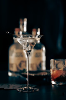 A sophisticated arrangement with a martini glass filled with a clear beverage topped with olives, accompanied by a glass of whiskey on the rocks and playing cards scattered on the table. In the background, blurred images of liquor bottles add an element of depth.