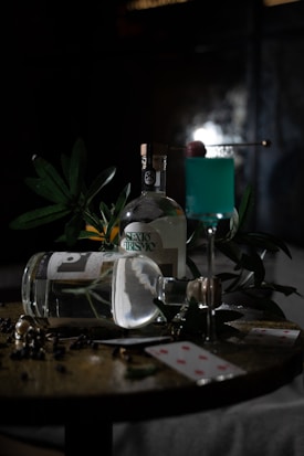 A dimly lit scene with a bottle and glass on a table, surrounded by foliage. The glass contains a blue cocktail with a red cherry garnish. Two playing cards are scattered across the surface, adding an element of intrigue and mystery.