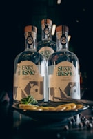 Three bottles labeled 'Sexto Abismo' with artistic designs on their labels are placed on a dark-colored surface. In front of the bottles, there is a plate with lemon slices and herbs.