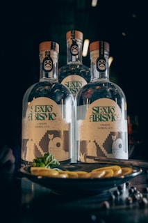 Three bottles labeled 'Sexto Abismo' with artistic designs on their labels are placed on a dark-colored surface. In front of the bottles, there is a plate with lemon slices and herbs.