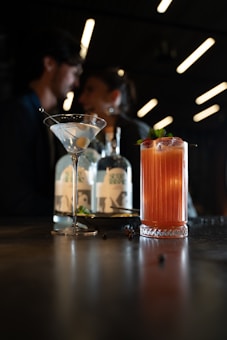 Two cocktails are placed on a dimly lit bar, one in a martini glass and another in a taller glass garnished with berries and mint. In the background, there are two blurred figures engaging in conversation, adding a sense of relaxed intimacy to the scene. Bottles of alcohol are also visible in the background.