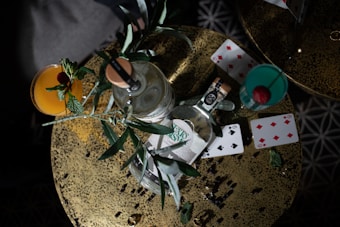 An overhead view of a table setup featuring two cocktails, a bottle of alcohol, playing cards, and some green leaves. The cocktails are garnished with fresh herbs and fruit, and the playing cards are scattered around the table, creating a casual and playful atmosphere.