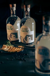 Two glass bottles with intricate labels stand on a dark surface. Slices of citrus fruit and a sprig of fresh mint rest on a plate nearby, accompanied by scattered small dark berries.