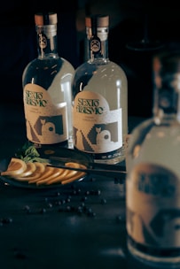 Two glass bottles with intricate labels stand on a dark surface. Slices of citrus fruit and a sprig of fresh mint rest on a plate nearby, accompanied by scattered small dark berries.