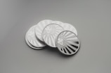 A collection of shiny silver coins with intricate engravings, placed on a smooth surface. Each coin features a detailed design of a lion's face surrounded by rays that resemble the sun.