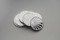 A collection of shiny silver coins with intricate engravings, placed on a smooth surface. Each coin features a detailed design of a lion's face surrounded by rays that resemble the sun.