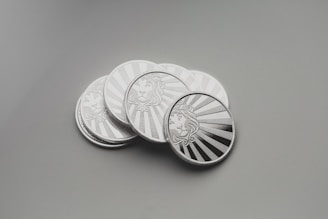 A collection of shiny silver coins with intricate engravings, placed on a smooth surface. Each coin features a detailed design of a lion's face surrounded by rays that resemble the sun.