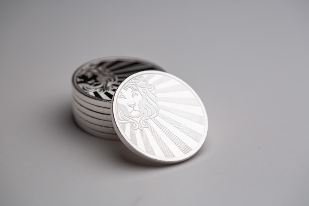 A stack of silver coins with intricate designs featuring a lion's head in the center against a backdrop of radiant lines. The coins are arranged neatly on a smooth, neutral-colored surface, creating a sense of order and precision.