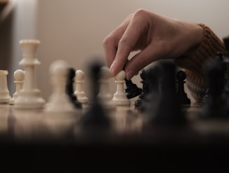 A hand is poised over a chessboard, gripping a white pawn while other chess pieces, both white and black, are arranged on the board. The image captures a moment of decision in the game, with a shallow depth of field focusing on the pieces and the hand.