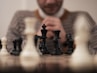 A person is sitting behind a chessboard, focusing on the game. The chess pieces are prominently displayed in the foreground with a combination of black and white pieces. The individual appears to be contemplating the next move, hands clasped together in front of their face.