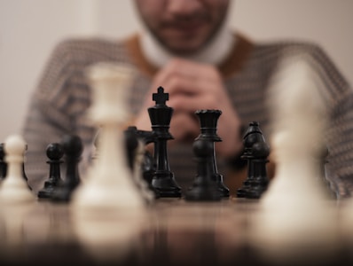 A person is sitting behind a chessboard, focusing on the game. The chess pieces are prominently displayed in the foreground with a combination of black and white pieces. The individual appears to be contemplating the next move, hands clasped together in front of their face.