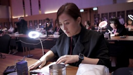 A woman is seated at a table with focused attention, working on a task under a bright circular lamp. She is surrounded by others engaged in similar activities within a room that appears to be a classroom or workshop.