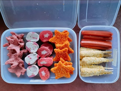 Various processed foods are arranged in two containers. On the left, there are pink, star-shaped pieces and round pink and white pieces with stars in the center. On the right, there are sausages and breaded skewers.