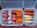 Various processed foods are arranged in two containers. On the left, there are pink, star-shaped pieces and round pink and white pieces with stars in the center. On the right, there are sausages and breaded skewers.