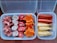 Various processed foods are arranged in two containers. On the left, there are pink, star-shaped pieces and round pink and white pieces with stars in the center. On the right, there are sausages and breaded skewers.
