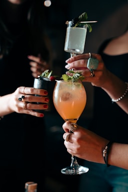 Several hands holding different types of cocktails; one glass is tall with a sprig of mint, another is in a rounded goblet with an orange-hued drink, and another is a short glass with a dark appearance and garnished with berries. The scene suggests a social gathering or celebration.
