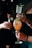Several hands holding different types of cocktails; one glass is tall with a sprig of mint, another is in a rounded goblet with an orange-hued drink, and another is a short glass with a dark appearance and garnished with berries. The scene suggests a social gathering or celebration.