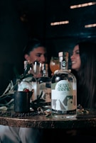 A wooden table displays a bottle labeled 'SEXO ABISMO', accompanied by a few symbolic objects like a mug with a raspberry and leaves, and a shiny ring. In the background, a blurred image of two people sits, suggesting a dimly lit, intimate setting with a convivial atmosphere.