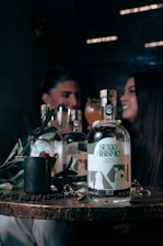 A wooden table displays a bottle labeled 'SEXO ABISMO', accompanied by a few symbolic objects like a mug with a raspberry and leaves, and a shiny ring. In the background, a blurred image of two people sits, suggesting a dimly lit, intimate setting with a convivial atmosphere.