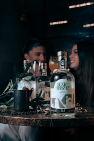 A wooden table displays a bottle labeled 'SEXO ABISMO', accompanied by a few symbolic objects like a mug with a raspberry and leaves, and a shiny ring. In the background, a blurred image of two people sits, suggesting a dimly lit, intimate setting with a convivial atmosphere.
