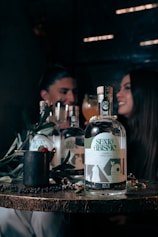 A wooden table displays a bottle labeled 'SEXO ABISMO', accompanied by a few symbolic objects like a mug with a raspberry and leaves, and a shiny ring. In the background, a blurred image of two people sits, suggesting a dimly lit, intimate setting with a convivial atmosphere.