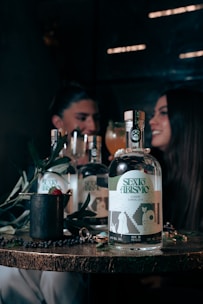 A wooden table displays a bottle labeled 'SEXO ABISMO', accompanied by a few symbolic objects like a mug with a raspberry and leaves, and a shiny ring. In the background, a blurred image of two people sits, suggesting a dimly lit, intimate setting with a convivial atmosphere.